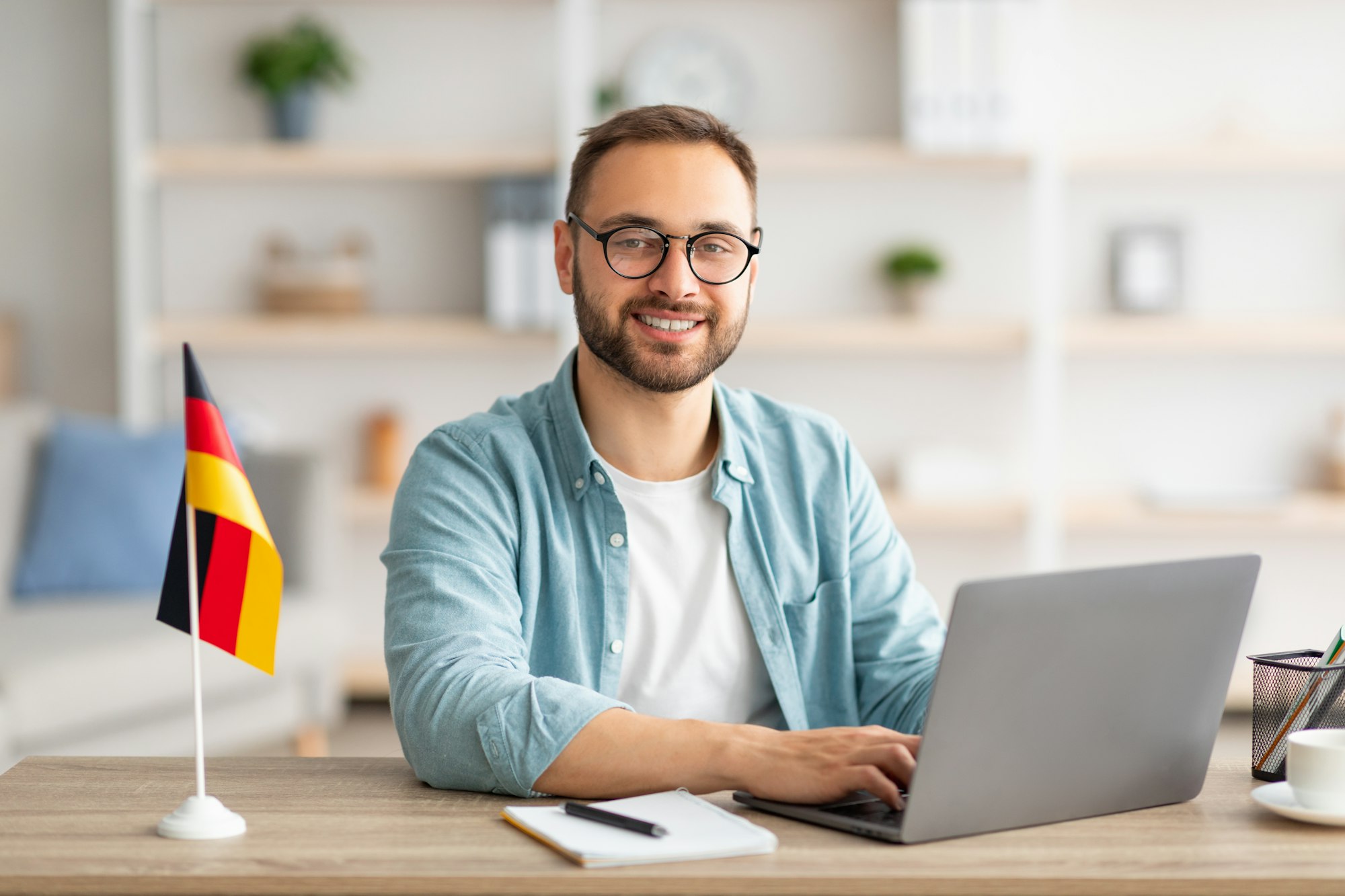 Happy young guy sitting at table with flag of Germany, using laptop pc, studying foreign language